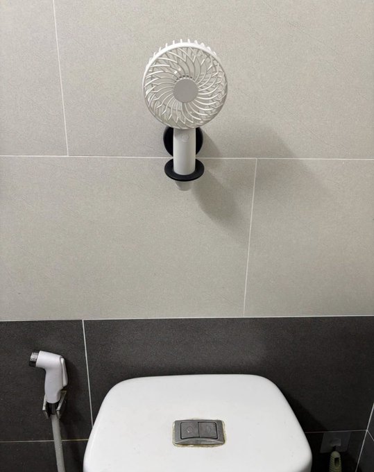 Bathroom scene featuring a white pedestal fan mounted on a black holder against a grey tiled wall, with the top of a white toilet tank and bidet sprayer visible below.