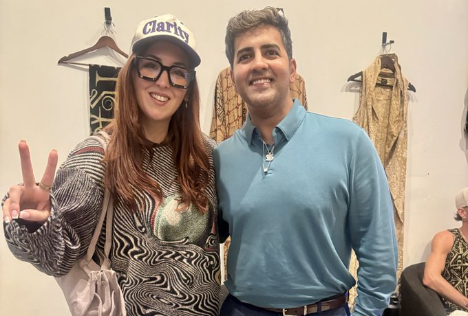 A selfie of a woman wearing a 'Clarity' cap and glasses making a peace sign, standing next to a smiling man in a blue polo shirt inside what appears to be a clothing store.