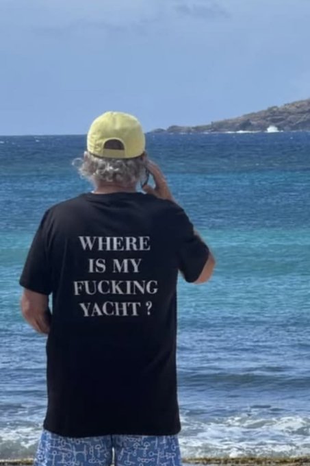 A man standing on a beach facing the ocean, wearing a black t-shirt with the text 'WHERE IS MY FUCKING YACHT?' and a yellow cap, holding his hand to his head.