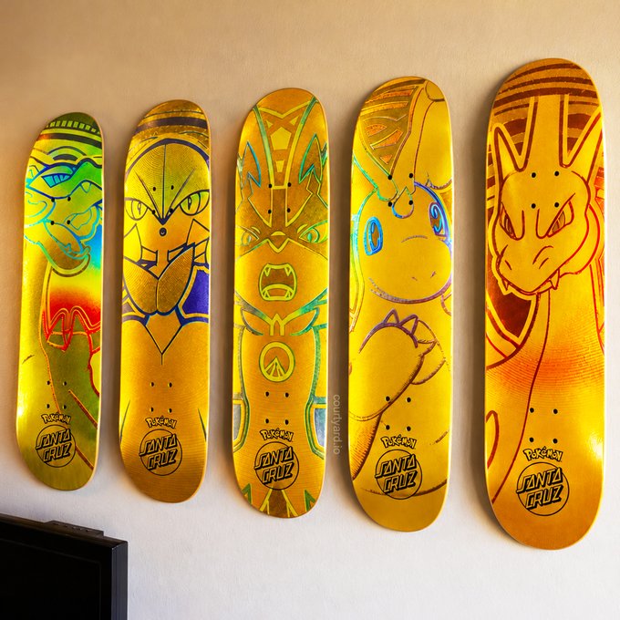 Five gold-colored Santa Cruz skateboards featuring Pokémon designs—including Bulbasaur, Lucario, Zeraora, Pikachu, and Charizard—displayed in a row mounted on a wall.