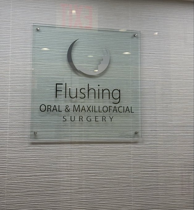A frosted glass office sign mounted on a textured wall that reads 'Flushing ORAL & MAXILLOFACIAL SURGERY' with a crescent moon logo.