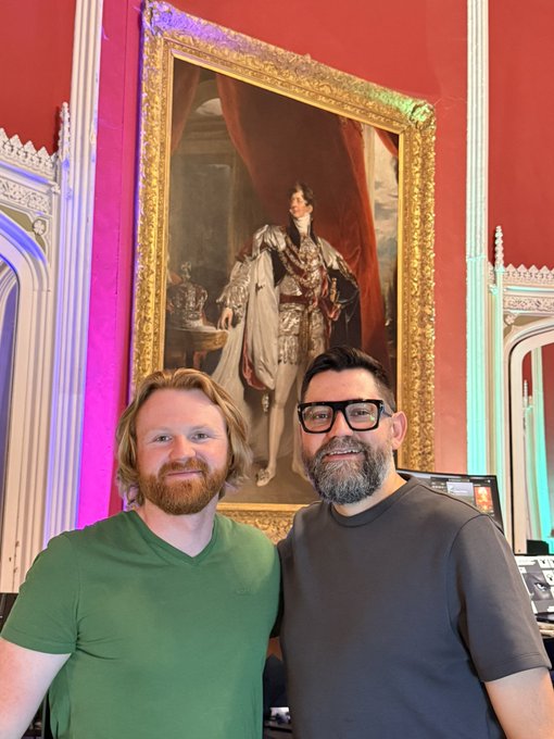 Two men smiling and posing for a photo in front of a large, ornate gold-framed portrait of a royal figure wearing a crown and ermine cape, with one man wearing a green shirt and the other a gray shirt and glasses.