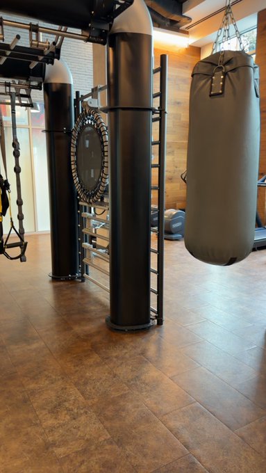 A home gym setup featuring two tall black freestanding heavy bags with integrated ladder rungs, suspended battle ropes, and a hanging punching bag on a tiled floor — ready for a "Gm" (good morning) workout session.