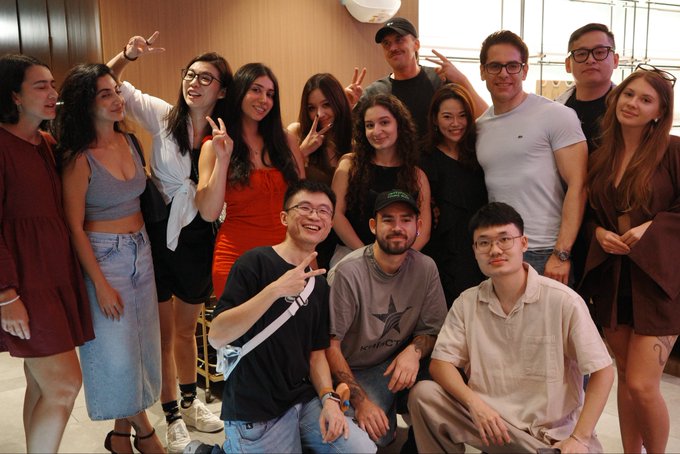 A group photo of around fourteen casually dressed young people smiling and posing with peace signs indoors.