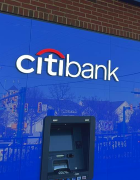 The blue exterior wall of a Citibank branch features the bank's white logo with a red arc above it, positioned above an ATM with reflections of buildings and trees visible in the glossy surface.