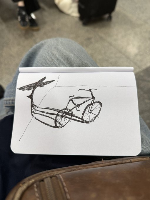 A pen sketch of a bicycle drawn in a small notebook resting on someone's denim-clad lap, illustrating the travel artwork shared on the artist's secondary Instagram account @ZanardiDraw.