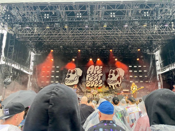 A concert photo taken from the audience showing a stage with skull graphics and a banner reading 'APE FEST 2022,' with band members performing and crowd members in rain ponchos visible in the foreground.