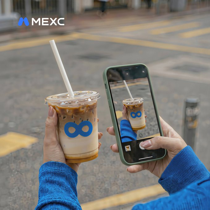 The image shows a person holding up their phone, taking a picture of a drink that has an "Infinite" symbol on it. The drink appears to be a coffee or iced beverage with a straw inside a cup. The person's reflection is visible in the camera lens, and they are using a blue cell phone for the photo. The image also contains text that reads: "No words. Just Show us what 'Infinite' looks like through your eyes: a photo, video, artwork or anything creative featuring the symbol. 200 $USDT Prize Pool for 10 winners: Follow @MEXC0fee Ends on May 31."