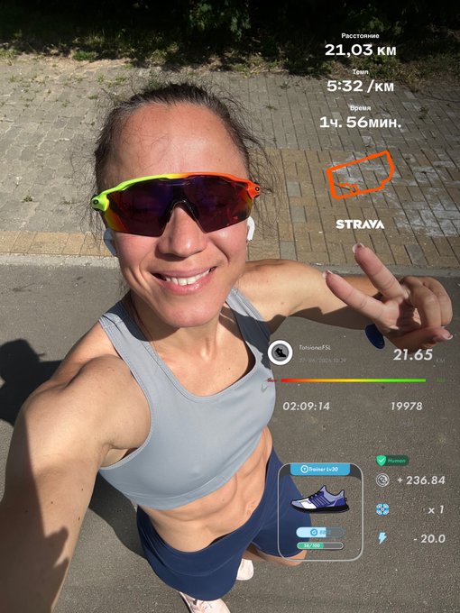 A smiling woman in a gray Nike sports bra and reflective sunglasses takes a selfie outdoors, flashing a peace sign after completing a 21.03 KM run with a 5:32/KM pace in 14:56 minutes, as tracked by the Strava app overlay on the image. The post celebrates her achievement while connecting it to her #STEPNathion fitness goal and the STEPN app, highlighted by the shoe NFT display showing +236.84 rewards.