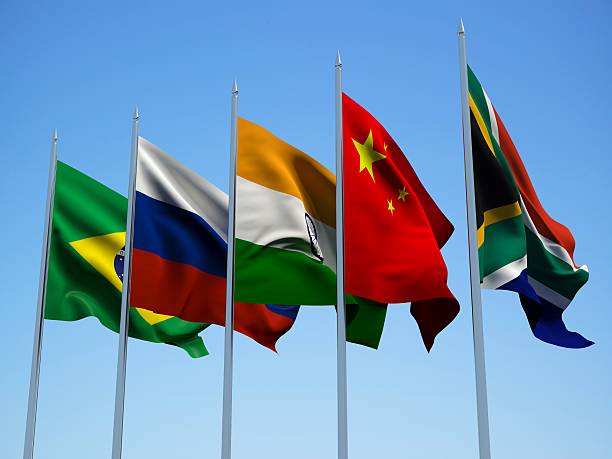The image features five flags from different countries fluttering in the wind against a clear blue sky. The flags are arranged in a line with each flag positioned at an angle relative to its neighbors. The text "HUGE: Ripple'S ODL expansion now spans Brazil, Mexico, India, and Southeast Asia." suggests that Ripple's new global cross-border payments market is expanding beyond the five countries featured in the image. This expansion could potentially connect various regions of the world through financial transactions, creating a network of commerce and exchange across borders.