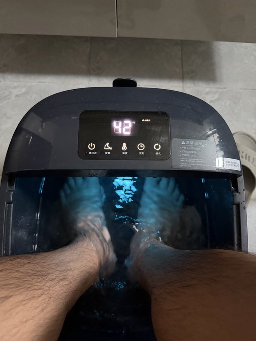 A pair of feet soaking in a foot bath/spa device with a digital display showing '98°' and Chinese warning text on the side.