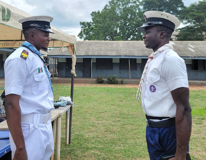Two men in white naval/military uniforms with peaked caps facing each other outdoors on a grassy area with a building and tent in the background.