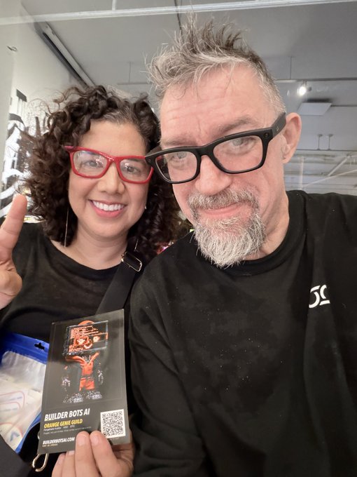 Two people, a woman with curly hair and red glasses, and a man with gray hair and black glasses, take a selfie holding a "BUILDER BOTS AI" badge labeled "ORANGE GENE GOLD."