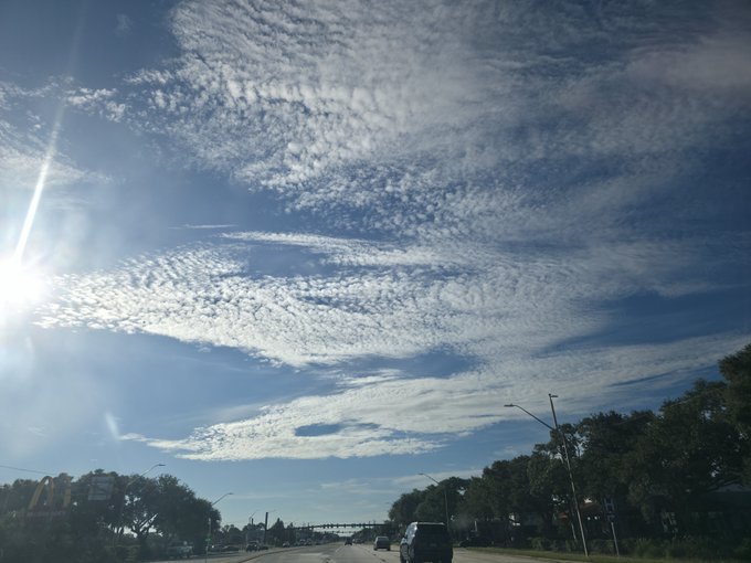 A daytime view from a car driving on a highway, showing a bright blue sky filled with textured altocumulus clouds and the sun shining brightly on the left.
