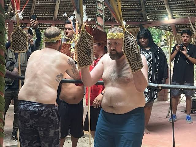 A gathering of indigenous people in traditional attire including feathered headdresses and woven shields, with two shirtless men holding decorated ceremonial items while others watch and photograph the event.