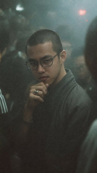 A man in glasses with a buzz cut and a ring gazes contemplatively at the camera in a dim, crowded setting, his thoughtful pose echoing the post's reflection on Pendle's rapid, understated rise as essential infrastructure across new chains like Monad, X Layer, and Robinhood Chain.