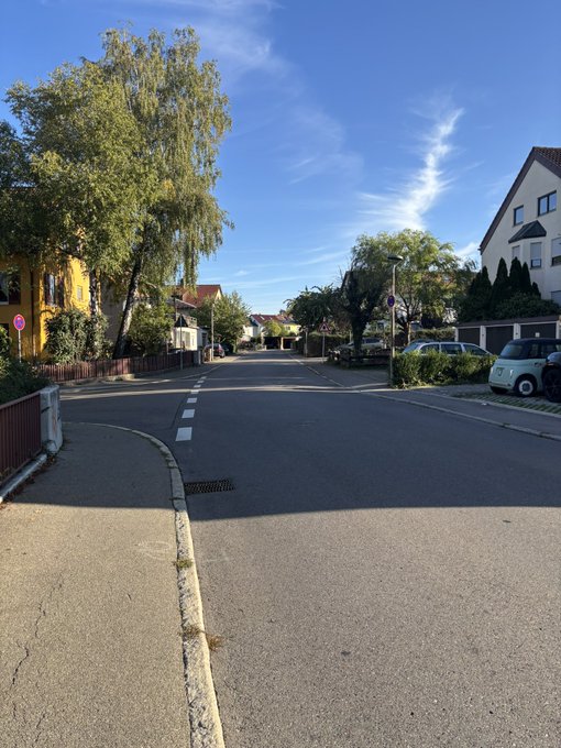 A quiet residential street lined with trees and houses under a blue sky with wispy clouds.