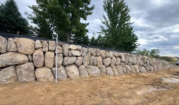 A stack of large boulders forms a sturdy stone wall, built from rocks sourced directly on-site to save thousands in material costs. A measuring pole in the foreground hints at the scale and craftsmanship of this Bitcoin-inspired "stack sats" project, achieved through trades and favors rather than cash.