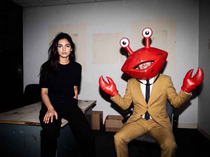 Two people pose in an office setting; a woman in black sits on a desk while a person in a tan suit wears a red crab head mask and matching red claw gloves.
