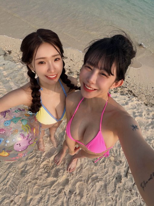 A selfie of two smiling women on a sandy beach, one in a yellow bikini and the other in a pink bikini with a palm tree tattoo on her arm, with a colorful inflatable swim ring visible in the background.