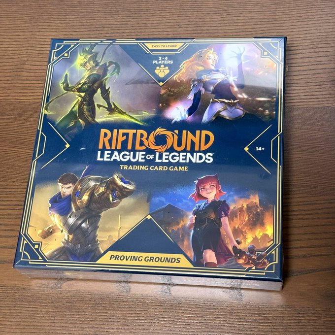 A box for the 'Riftbound: League of Legends Trading Card Game - Proving Grounds' set sits on a wooden surface, featuring artwork of four champions and noting it is for 3-4 players, easy to learn, and ages 14+.