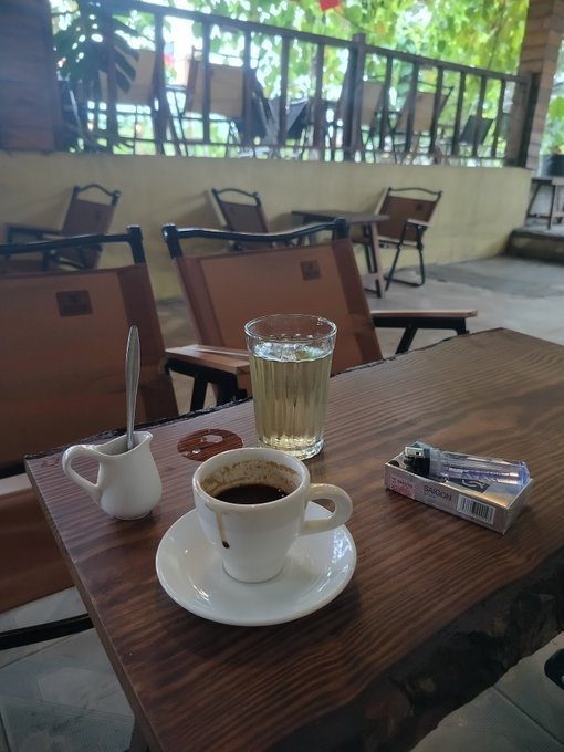 A small cup of dark coffee and a glass of water sit on a wooden table at an outdoor cafe, with empty chairs and tropical greenery visible in the background.
