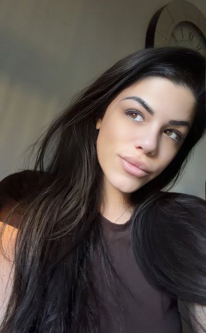 A young woman with long dark hair and a brown top poses for a close-up selfie, looking slightly upward with a soft expression.