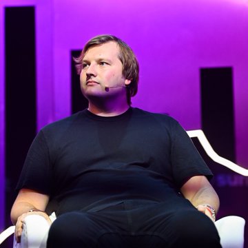 A man wearing a black t-shirt and headset microphone sits on a white chair against a purple-lit stage background, appearing to be speaking at a conference or panel discussion. The image accompanies a quote from Aave founder Stani Kulechov discussing the future regulatory path of decentralized finance (DeFi), likely depicting him during a speaking engagement on the topic.