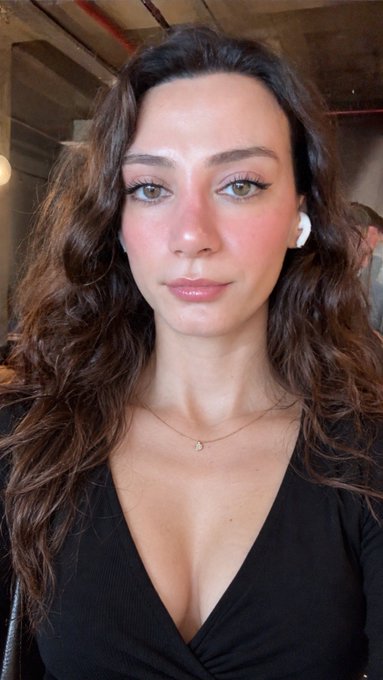 A portrait of a woman with long, wavy dark hair, wearing a black top and a delicate gold necklace with a small pendant. She has subtle makeup and is wearing white wireless earbuds.