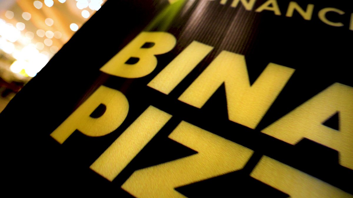 The image shows a close-up of a sign for a pizza restaurant called Bin Pizza. The sign has yellow letters that stand out against the black background. The text "Bin Pizza" is prominently displayed, along with the words "Pizza" and "Lahoris For". The sign also includes some numbers and a logo, possibly representing the restaurant's financial information or branding.