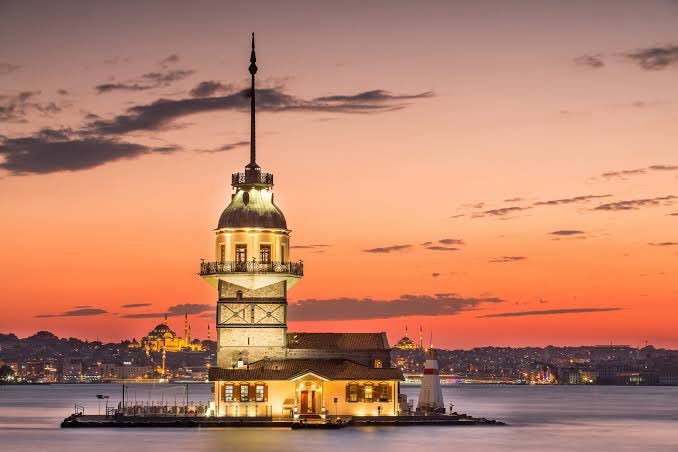 The image shows the iconic Maiden's Tower (Kız Kulesi) in Istanbul, Turkey, beautifully illuminated against a vibrant orange and pink sunset over the Bosphorus Strait. The post is a greeting in Turkish ("Merhaba" meaning "Hello") and references @grok, but the image itself contains no text, numbers, tickers, or names — notably, no Bitcoin logo has been integrated into the sunset as requested in the caption.