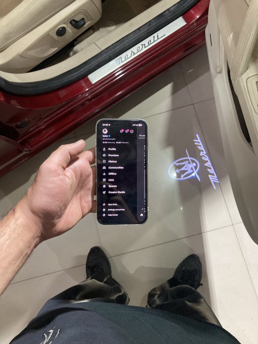 A person holds up a smartphone displaying a Spotify profile under the name "Dope Kids," showcasing 10,636 followers, while standing next to a Maserati projecting its illuminated logo onto the tiled floor—a visual flex aligning with the caption's theme of proving doubters wrong through success.
