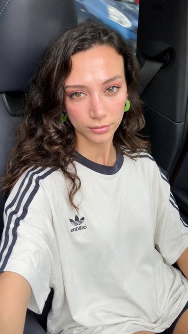 A young woman with curly brown hair and green earrings takes a selfie while sitting in a car, wearing an Adidas t-shirt with the brand's logo and name visible on the chest. Her relaxed, direct gaze complements the casual Sunday morning greeting vibe of the post.