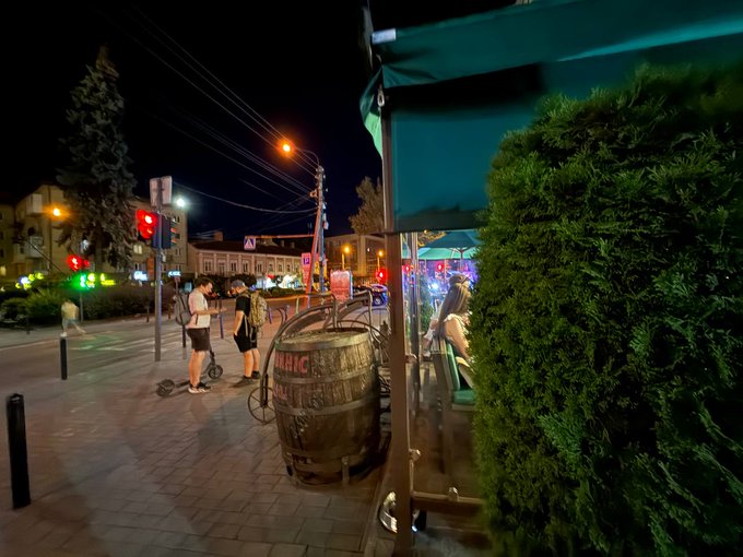 Nighttime street scene featuring a wooden barrel on a sidewalk, pedestrians walking, traffic lights, and a greeeen awning in the foreground.