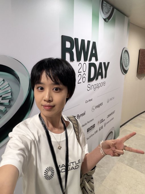 A selfie of a young woman with short black hair wearing a white 'Manila Squad' t-shirt and lanyard, posing in front of a banner for 'RWA Day 2026 Singapore' while making a peace sign.