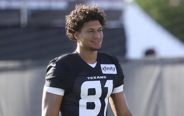 A smiling young man wearing a Houston Texans jersey with the number 81 and an "Xfinity" sponsor patch stands on the practice field. The image relates to the post about Texans wide receiver Jayden Higgins' torn ACL injury suffered during a joint practice against the Raiders.