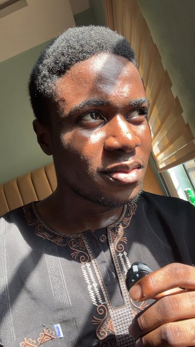A close-up portrait of a young Black man with short hair and a beard, wearing a dark embroidered traditional-style shirt, holding what appears to be an earbud case, with strong natural lighting on his face.