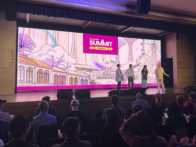 A photograph of a conference stage at the "IDG SUMMIT Singapore," where four presenters stand before a large illustrated backdrop; the audience is visible in the foreground watching the presentation.