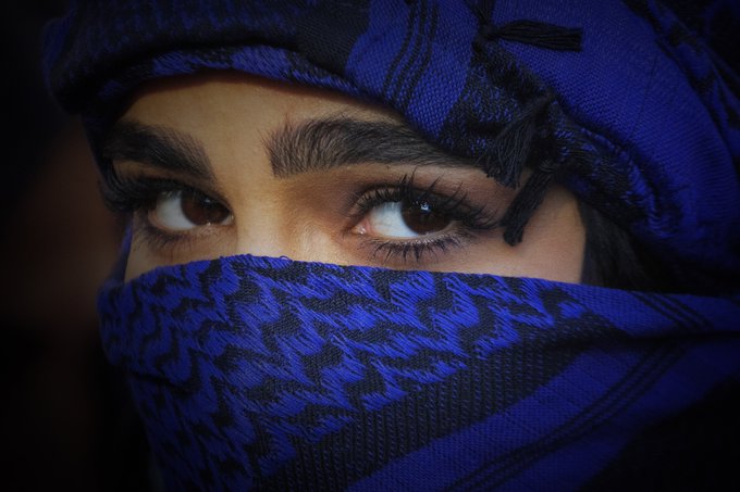 A close-up of a person's eyes and eyebrows, with the lower half of their face covered by a blue patterned scarf and head wrap.