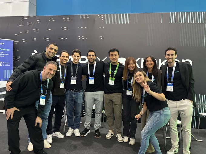 A group photo of ten smiling people wearing lanyards and mostly black attire, posing together in front of a black wall with white dotted patterns at what appears to be a corporate event or conference.
