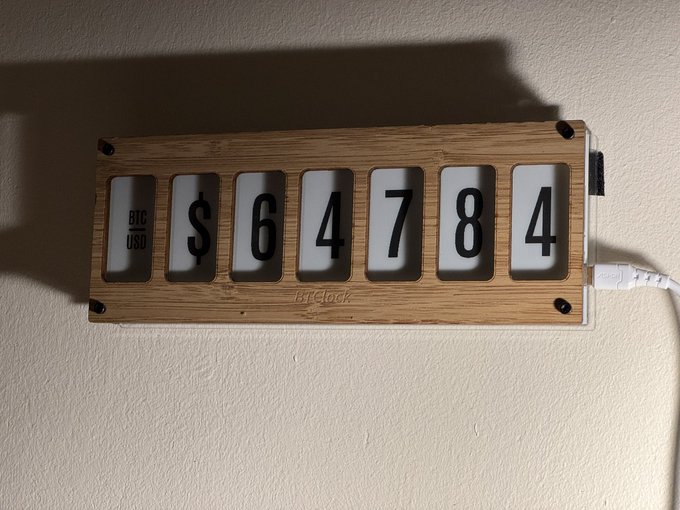 A wooden wall-mounted device by 'btClock' displays the Bitcoin price as '$64784' in large mechanical flip-style numbers, with a USB power cable plugged into its side.