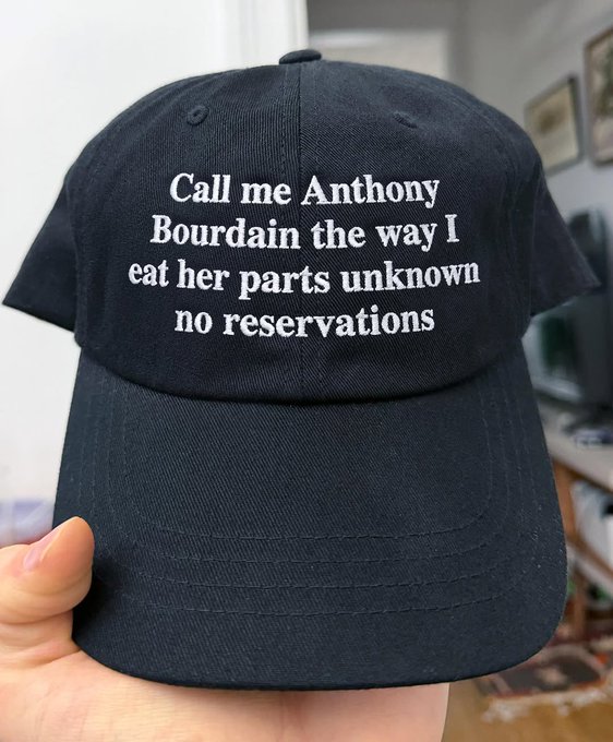 A black baseball cap is held up, featuring white embroidered text that reads: "Call me Anthony Bourdain the way I eat her parts unknown no reservations."