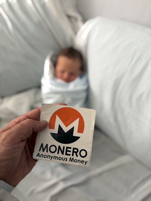 A hand holding a Monero ('Anonymous Money') sticker in front of a swaddled baby on a couch.
