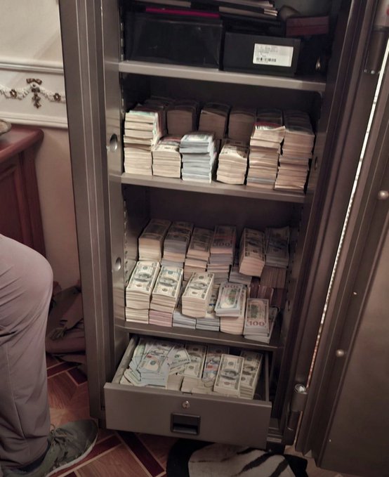 An open metal safe filled with numerous stacks of U.S. hundred-dollar bills arranged on multiple shelves.