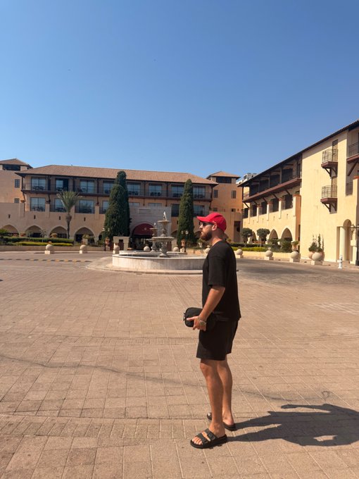 A man in a red cap, black t-shirt, and shorts stands in a sunlit courtyard holding a camera, with a fountain and Mediterranean-style buildings in the background.