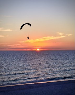 A serene sunset beach scene featuring a parasail silhouetted against the orange sky over the ocean.