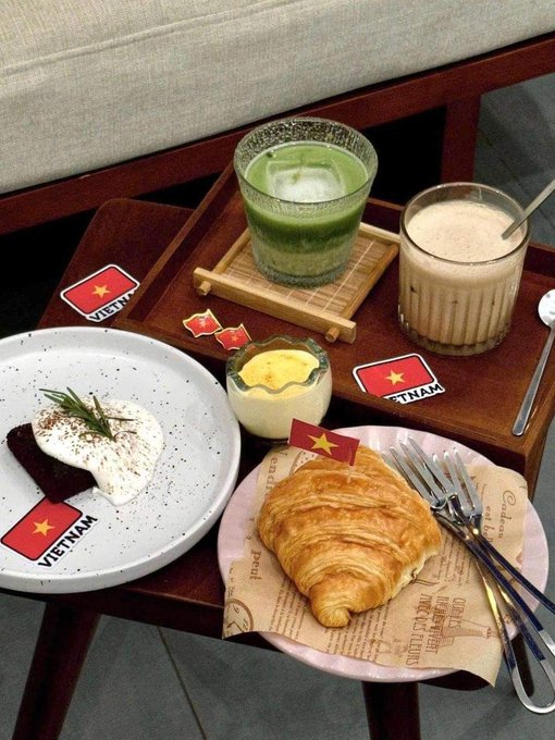 A Vietnamese-themed breakfast spread features a matcha latte, a coffee beverage, a chocolate brownie with cream and rosemary garnish, a crème brûlée, and a croissant on a wooden table decorated with multiple "VIETNAM" flag stickers. The leisurely café meal reflects the post's relaxed "Good Morning" mindset of enjoying a holiday break before diving into Monday's work.