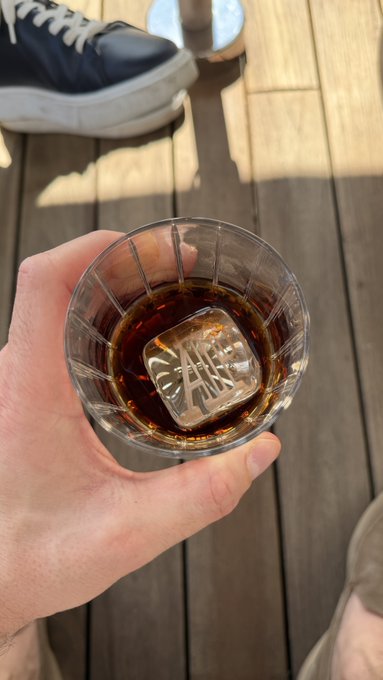 A hand holds a glass of what appears to be whiskey with a large ice cube engraved with "JANE DOE," raised in a casual toast on a wooden deck, paired with a navy sneaker in the background—perfectly capturing a relaxed "GM frens and happy day!" vibe.