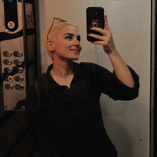 Mirror selfie of a woman with short blonde hair and sunglasses on her head taking a photo in an elevator with her phone.
