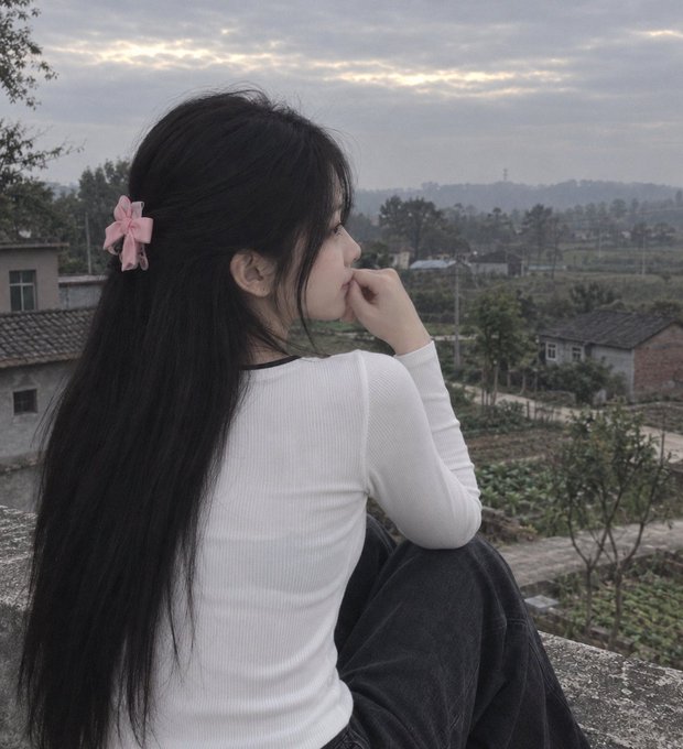 A young woman with long black hair tied with a pink bow sits on a ledge overlooking a rural village at dusk.